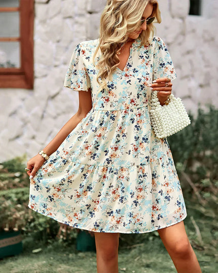 Women's cream floral print mini sundress with v-neck, flutter sleeves, and tiered skirt, holding a pearl bag.