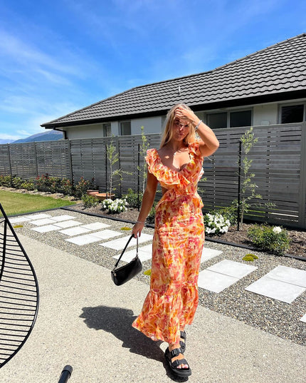 Full-length view of a women's floral ruffle tie-back maxi sundress in orange and pink, paired with black slides.