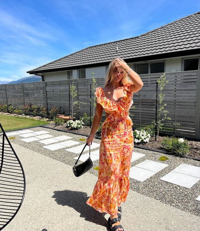 Full-length view of a women's floral ruffle tie-back maxi sundress in orange and pink, paired with black slides.