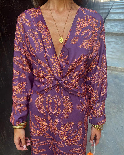 Close-up of a women's purple V-neck midi dress with an orange abstract print, twisted front detail, and gold necklace.