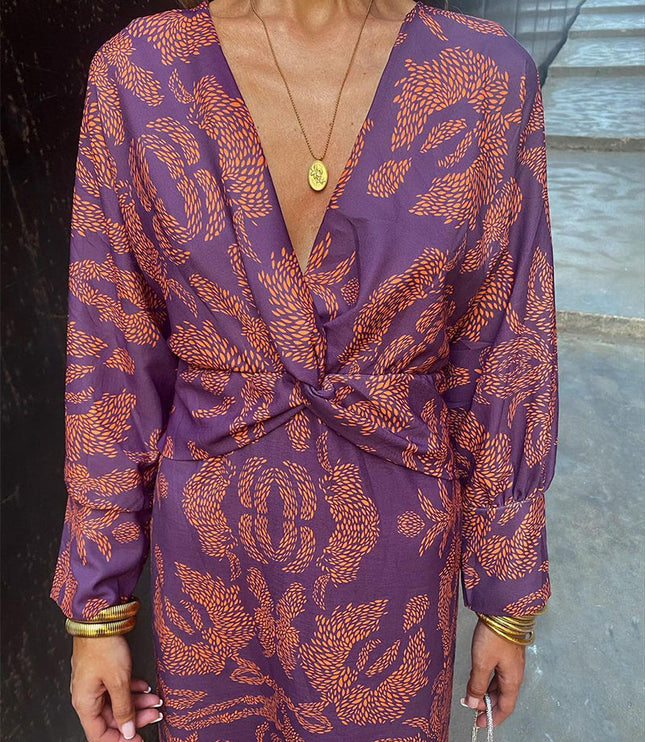 Close-up of a women's purple V-neck midi dress with an orange abstract print, twisted front detail, and gold necklace.