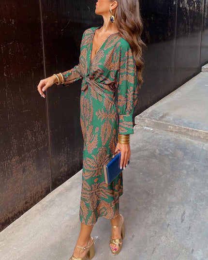 Women's summer midi dress in emerald green with an orange abstract print, V-neck, and long sleeves, styled with gold heels and a blue clutch.