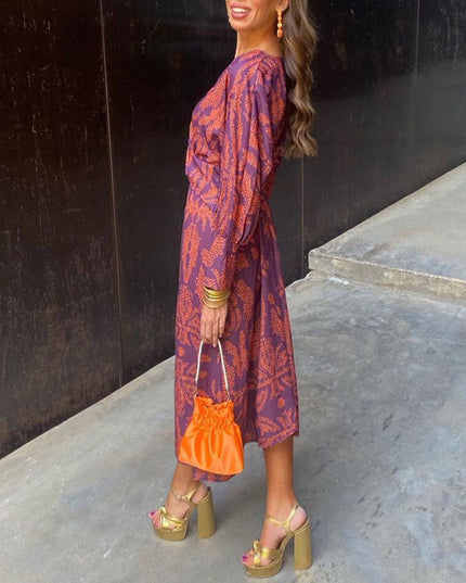 Side view of a women's purple summer midi dress with an orange abstract print, long sleeves, paired with gold platform heels and an orange purse.