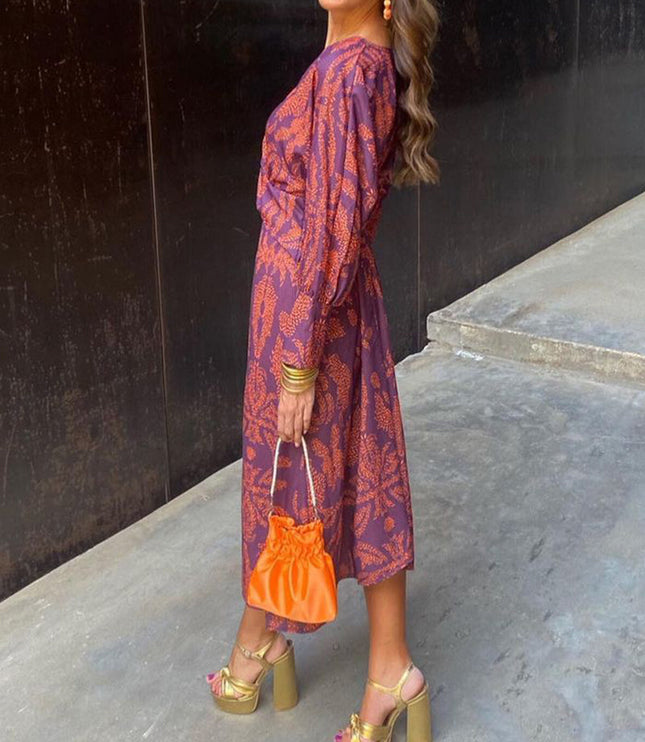 Side view of a women's purple summer midi dress with an orange abstract print, long sleeves, paired with gold platform heels and an orange purse.