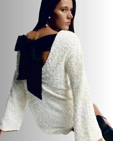 White textured mini dress with an open back and oversized black bow, shown in a seated pose
