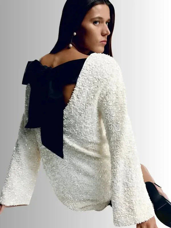 White textured mini dress with an open back and oversized black bow, shown in a seated pose