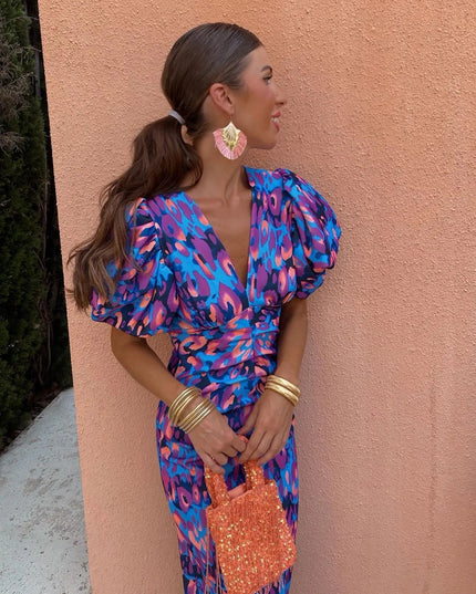 Women's cocktail dress in a blue and purple floral pattern with puff sleeves, V-neck, and cinched waist, styled with gold bangles.