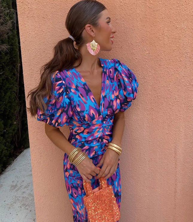 Women's cocktail dress in a blue and purple floral pattern with puff sleeves, V-neck, and cinched waist, styled with gold bangles.