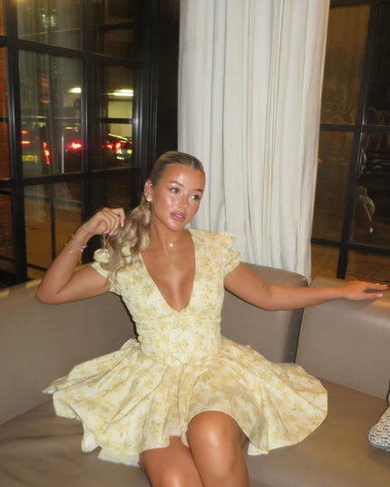 Yellow floral V neck mini dress with puff sleeves and gathered waist shown in a seated pose